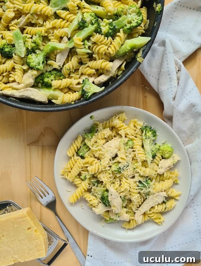 A plate of cooked chicken and broccoli pasta with freshly grated parmesan cheese on top. A fork on the left and a loosely folded kitchen towel on the right side of the plate.  A cooked chicken and broccoli pasta inside a large pan/skillet on the op left hand corner of image.  