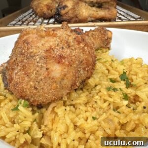 A crispy, golden-brown baked chicken drumstick sits atop a bed of vibrant Lemon Pepper Rice, garnished with herbs, in a white bowl. In the background, more chicken rests on a cooling rack.
