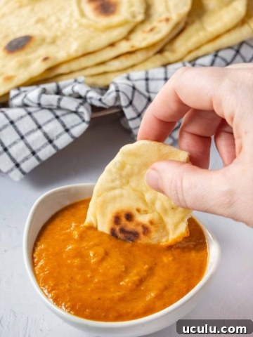 A hand dipping piece of naan bread into a small white bowl of orange sauce.