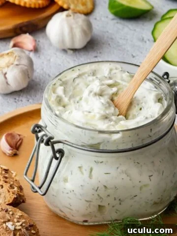 Creamy herbed white sauce in a jar surrounded by garlic, cucumbers, bread and crackers.