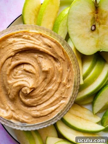 A glass bowl with a creamy brown dip surrounded by sliced apple.