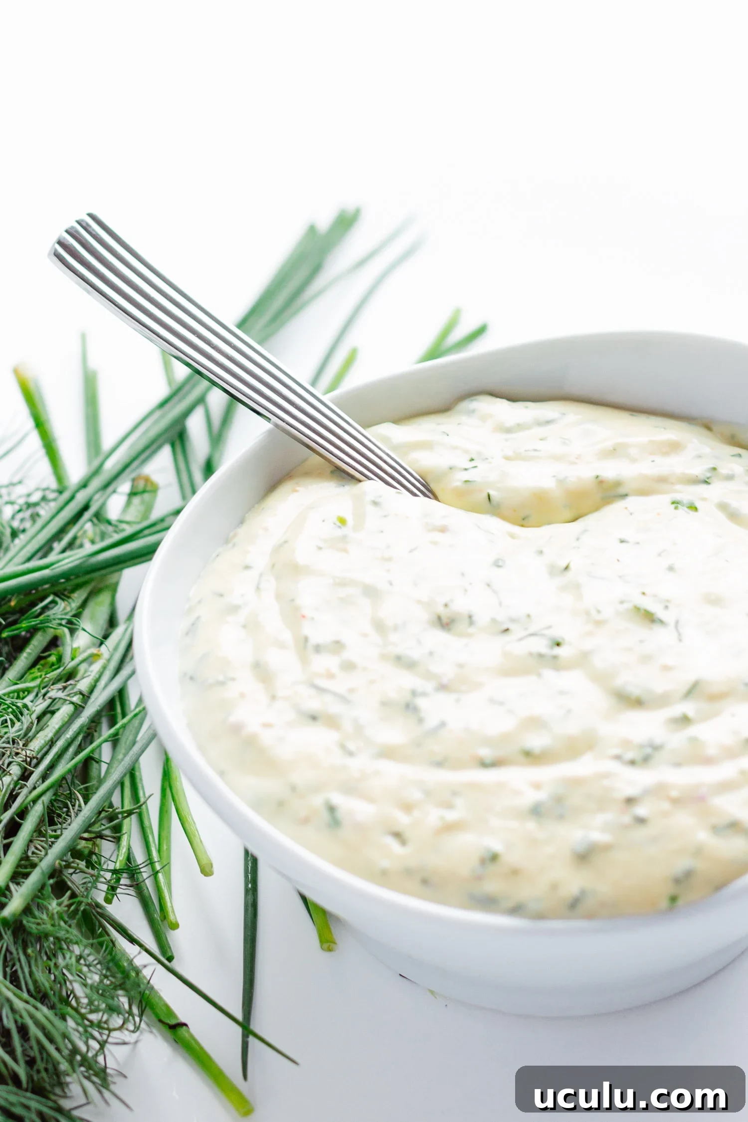 White bowl of creamy herb sauce/dip with spoon handle sticking out.  Scattering of dill and chives on the side.