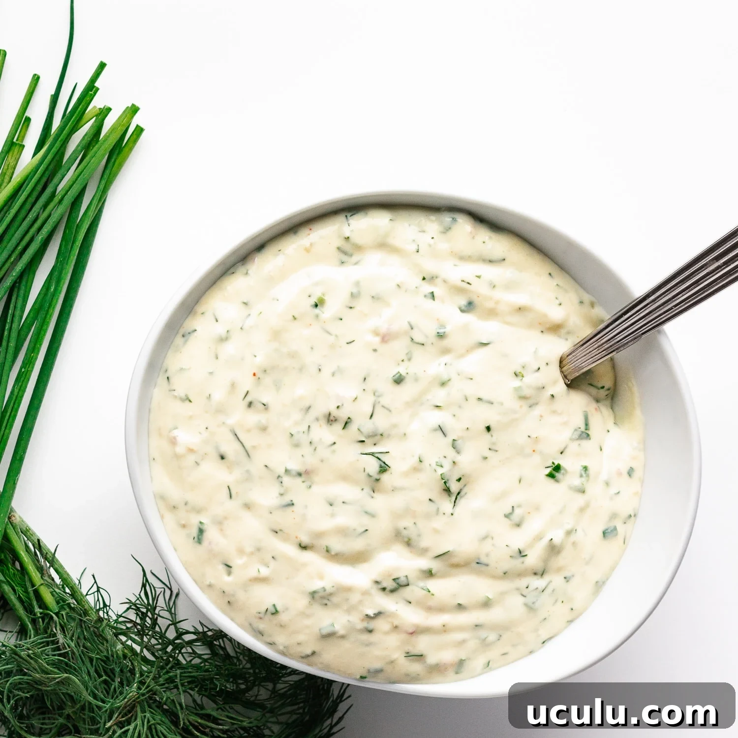 Overhead photo of creamy herb sauce/dip in a white bowl with silver spoon handle sticking out.  Scattering of chives and dill to the left of the bowl.