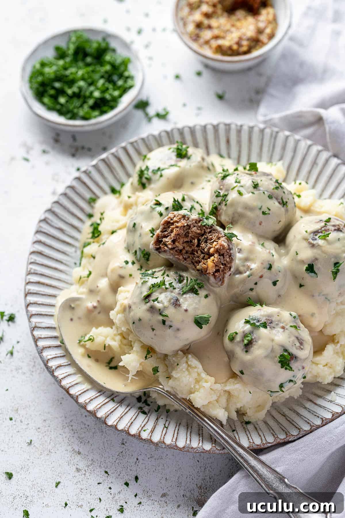 Vegan Swedish meatballs and mashed potato on a white plate with a bowl of parsley and a bowl of mustard.