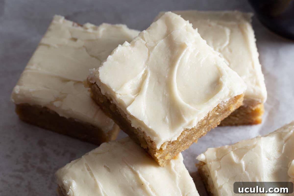 A group of blondie brownies that have been frosted with cream cheese icing sitting on piece of parchment paper.
