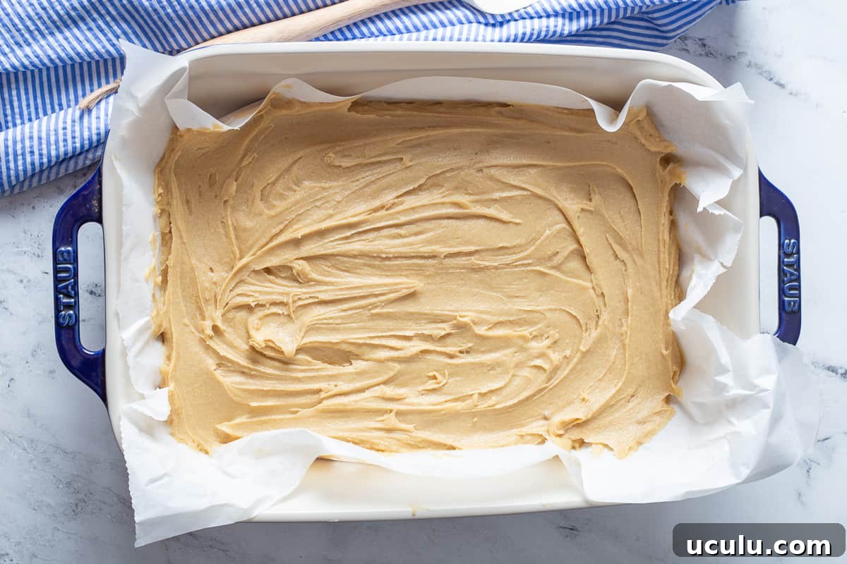 Batter for blondies is spread in 9x13 inch parchment paper lined baking dish.