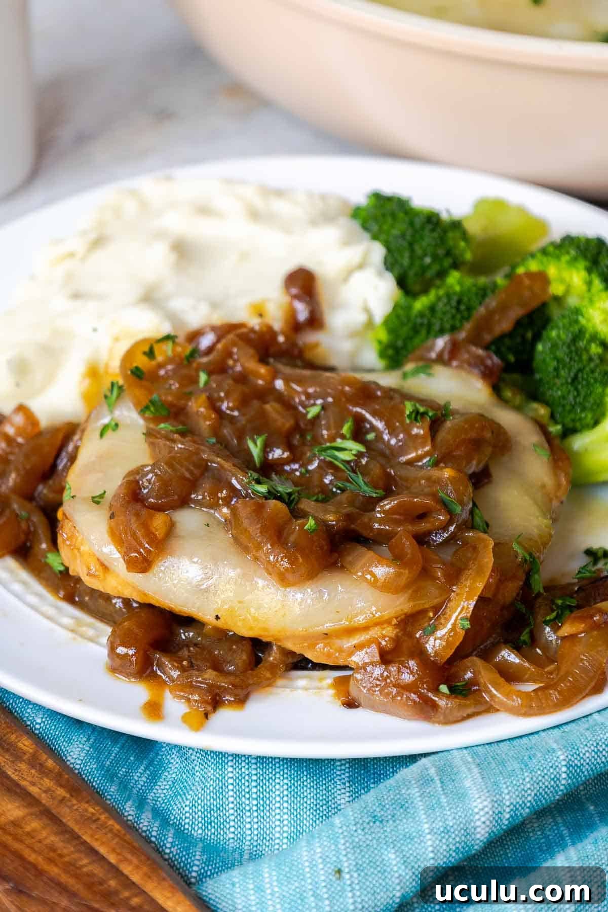 A pork chop on a plate covered in caramelized onions and cheese with mashed potatoes and broccoli.