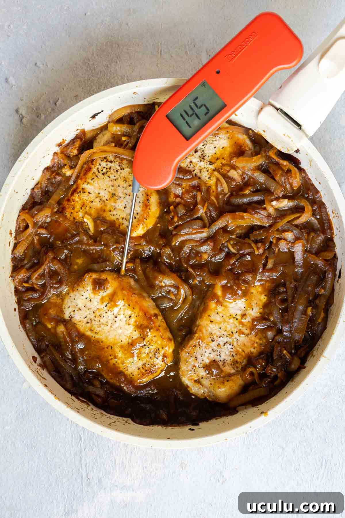 A meat thermometer in a pork chop showing 145°F, with the chops simmering in caramelized onions.