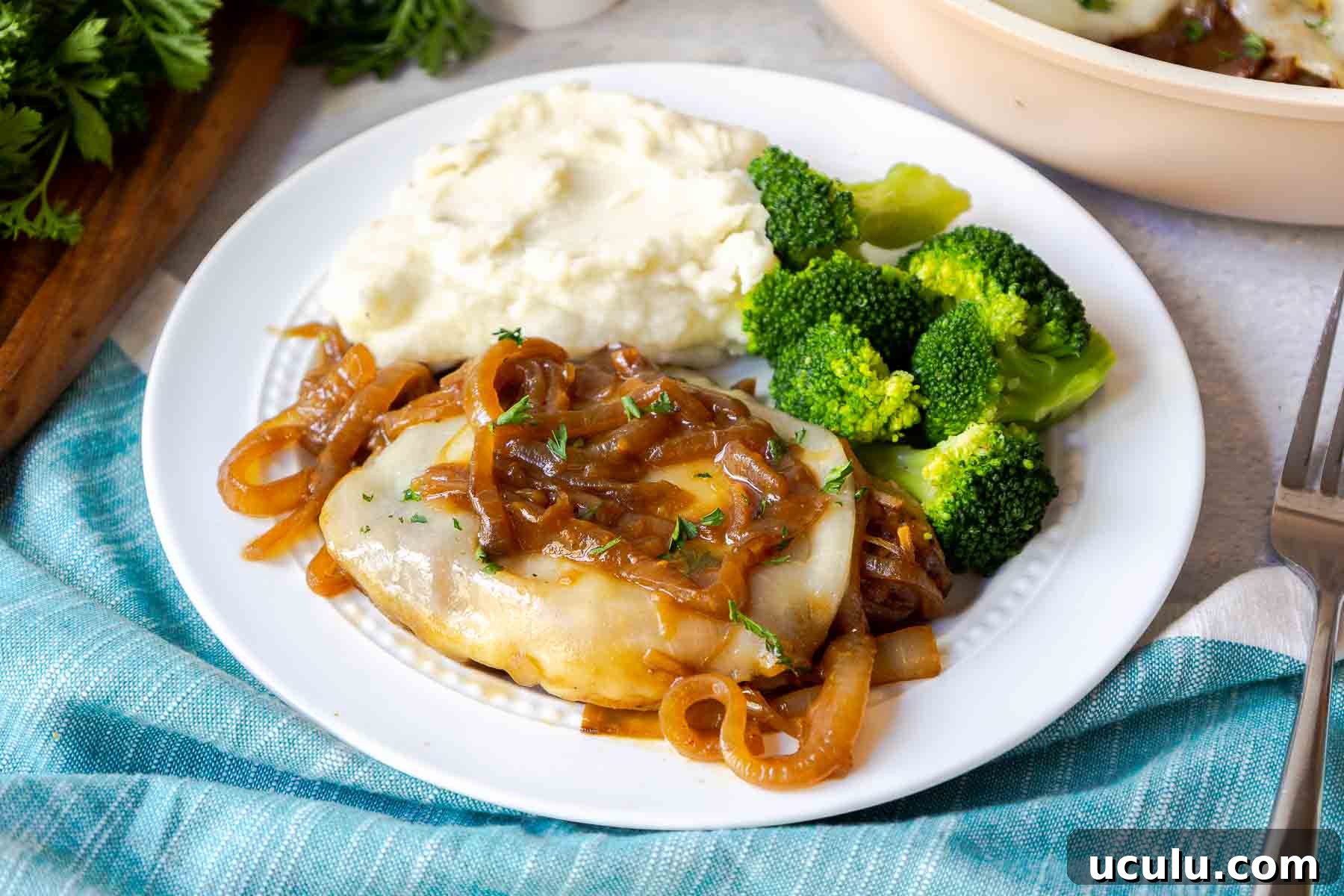 A pork chop topped with onions and cheese on a plate with mashed potatoes and broccoli.
