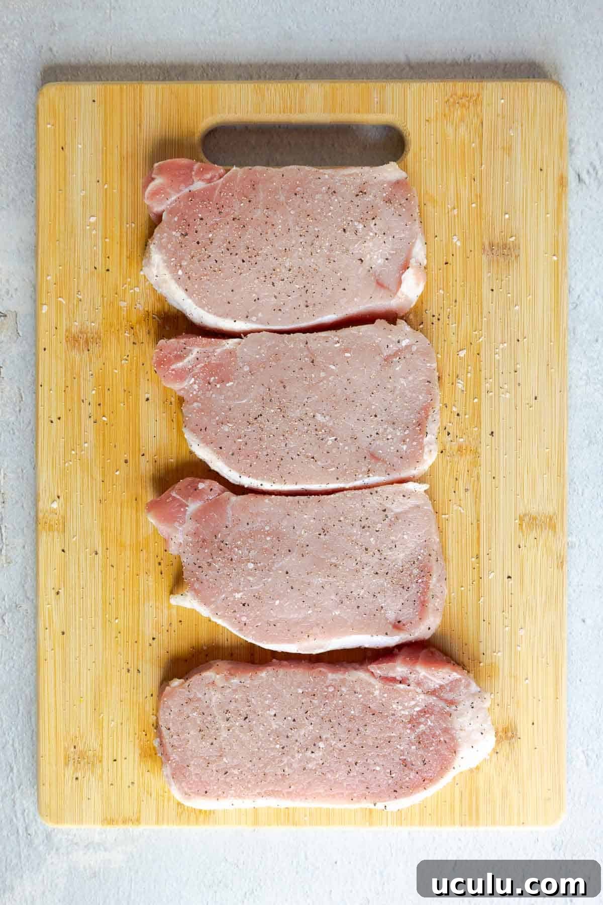 Four raw pork chops seasoned with salt and pepper on a wooden cutting board.