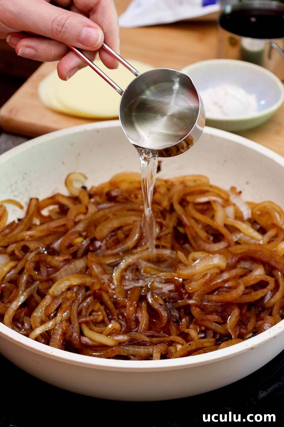 A hand pouring liquid from a measuring cup into a skillet of caramelized onions.