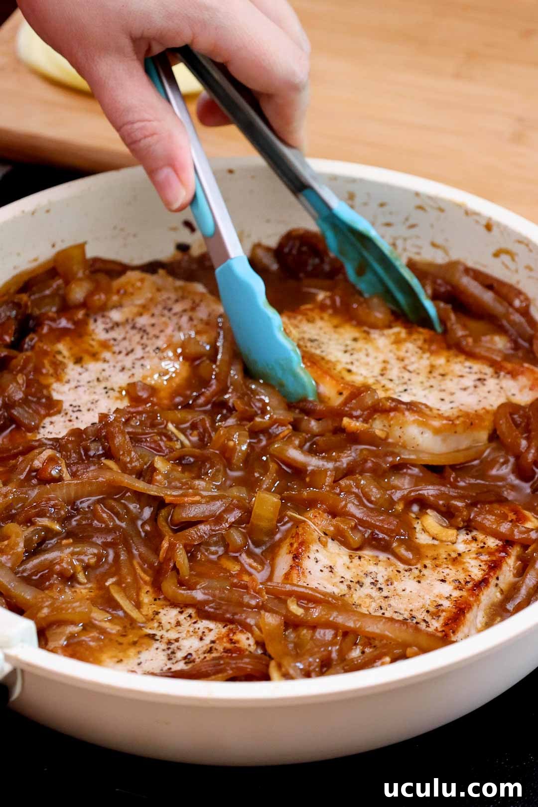 Tongs placing pork chops into a skillet with caramelized onion sauce.