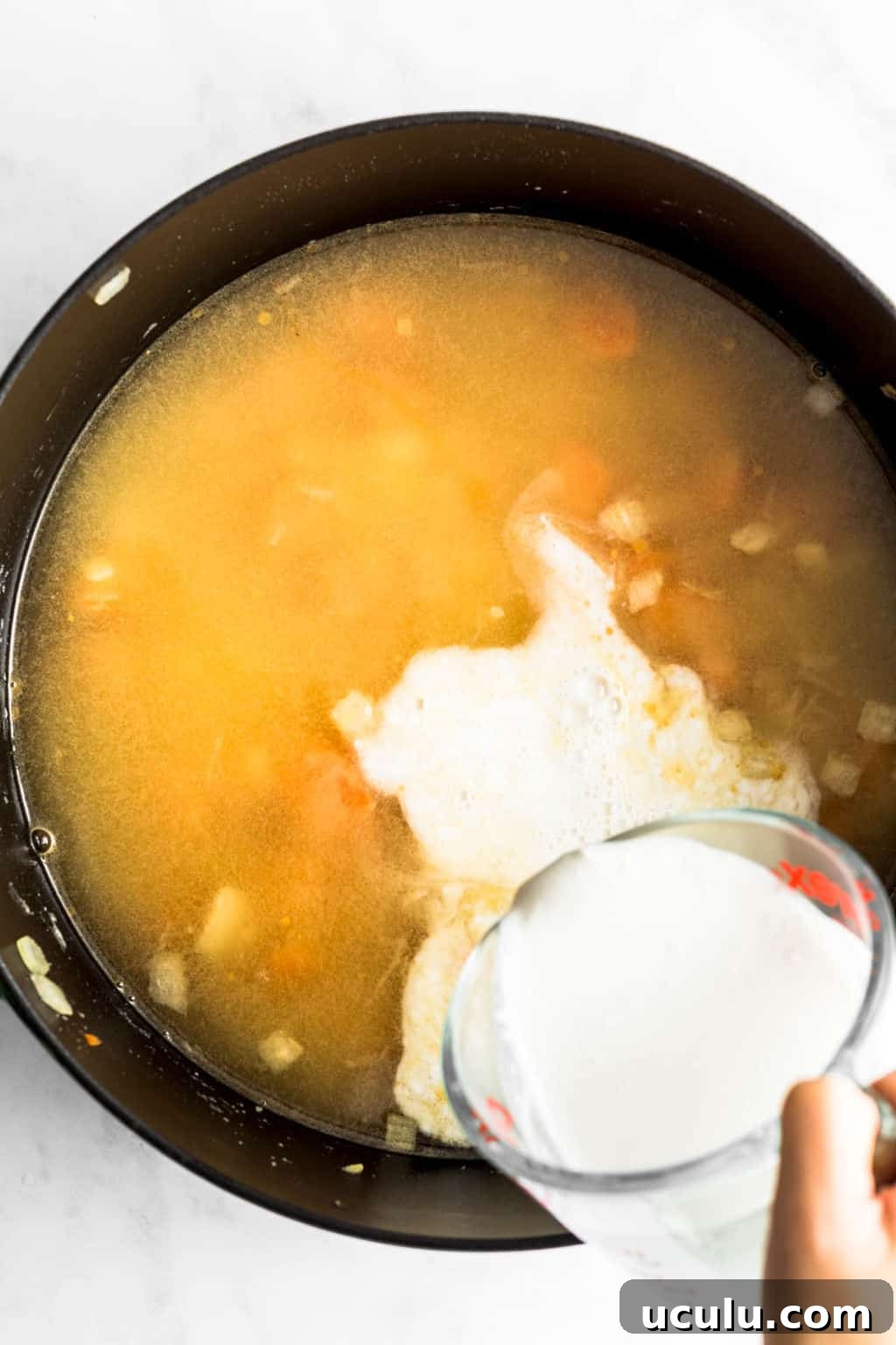 womans hand pouring white cream into a black pot filled with broth.