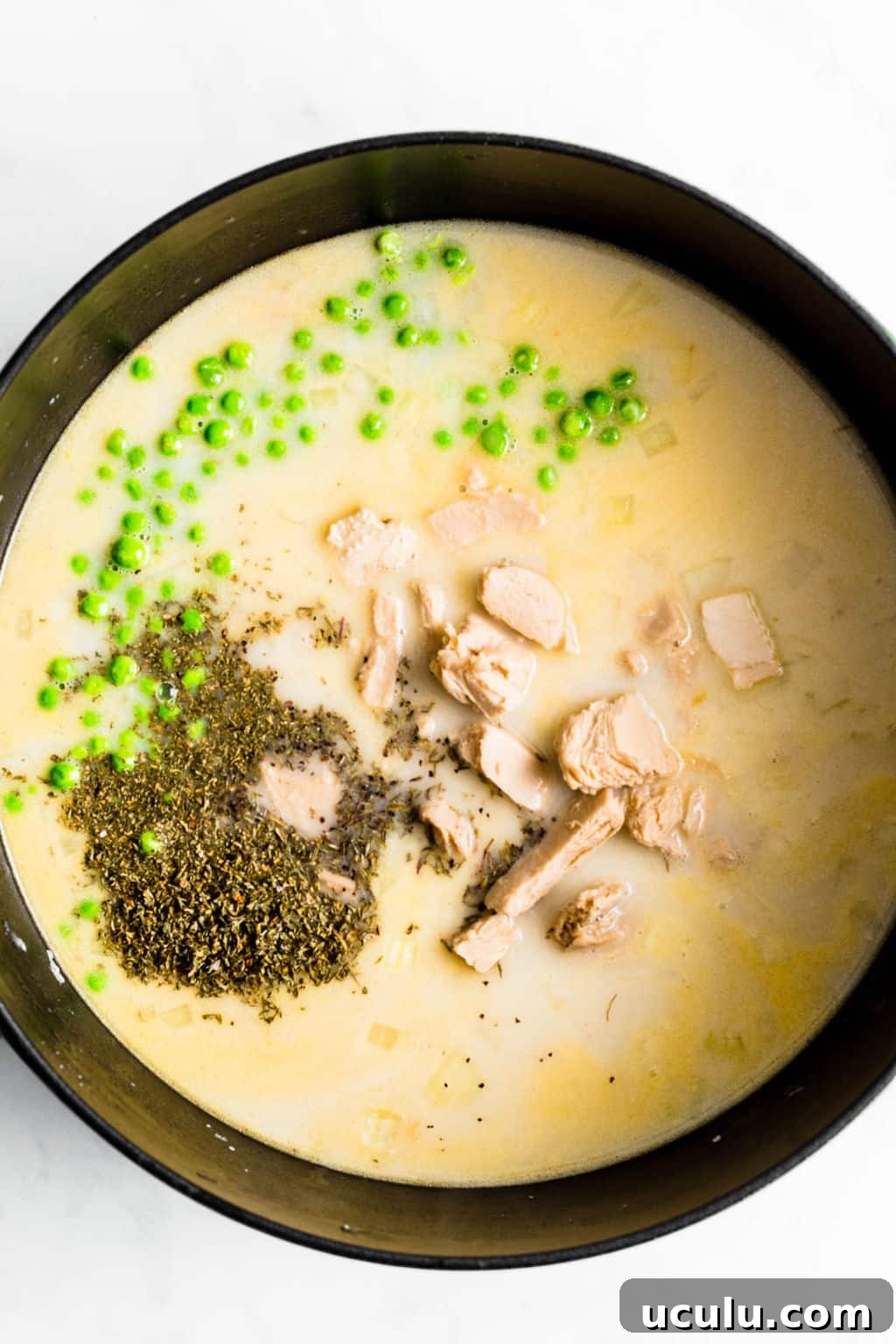 large black pot filled with a creamy liquid, vegan chicken, dried herbs, and green peas.