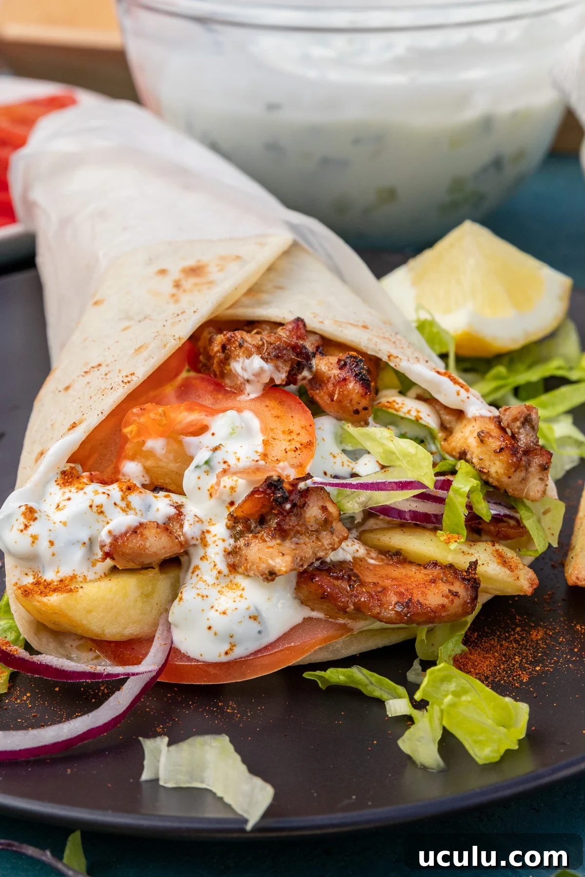 closeup of the inside of a Greek chicken gryo wrap with chicken thigh, tzatziki, onion, tomatoes and lettuce on a black plate with a bowl of tzatziki in the background.