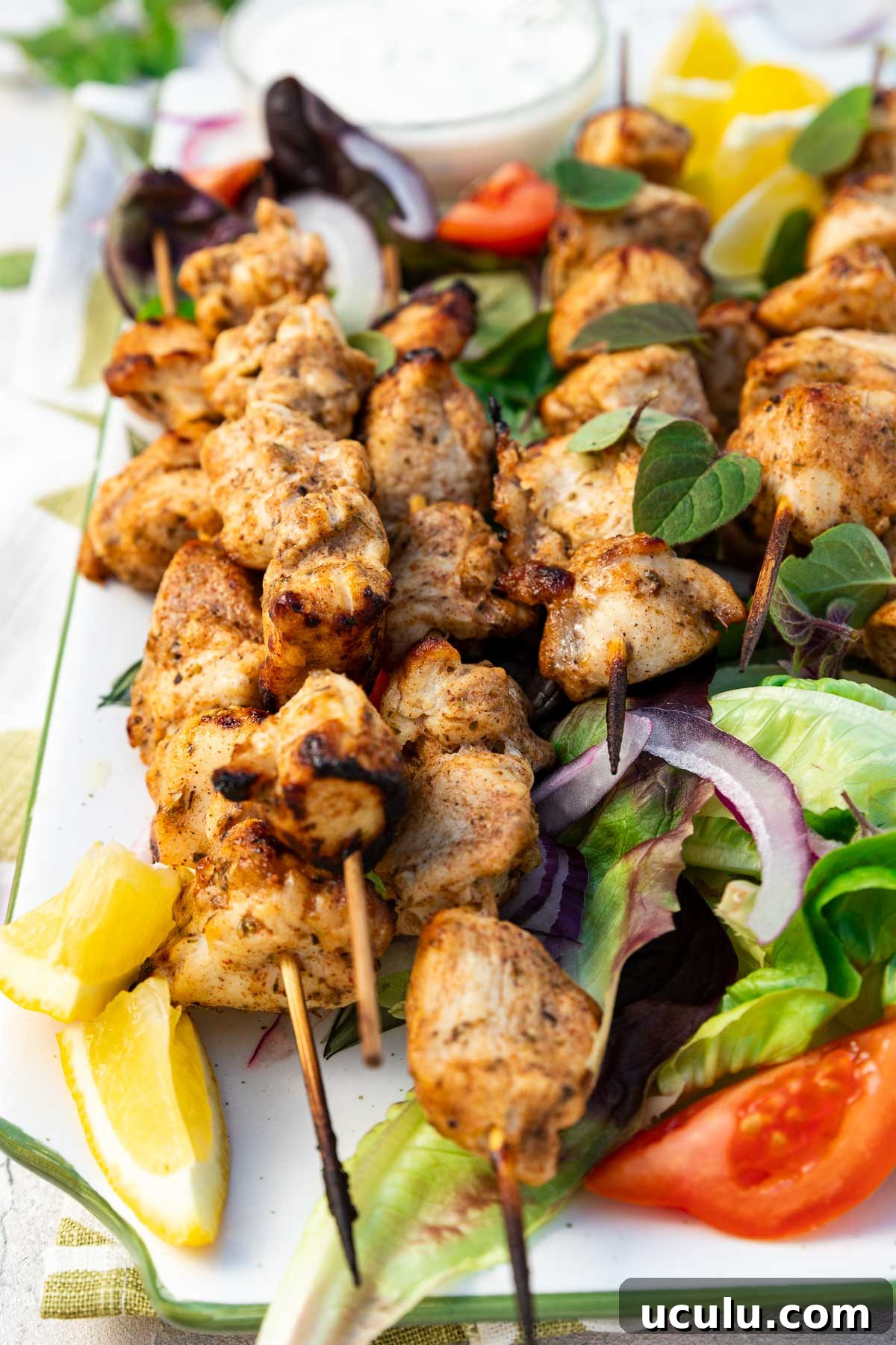 Closeup of a rectangular platter of baked chicken kabobs on a bed of simple salad and with lemon segments and a white dip on a green and white tea towel and on a pale textured background.