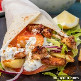 closeup of the inside of a Greek chicken gryo wrap with chicken thigh, tzatziki, onion, tomatoes and lettuce on a black plate with a bowl of tzatziki in the background.
