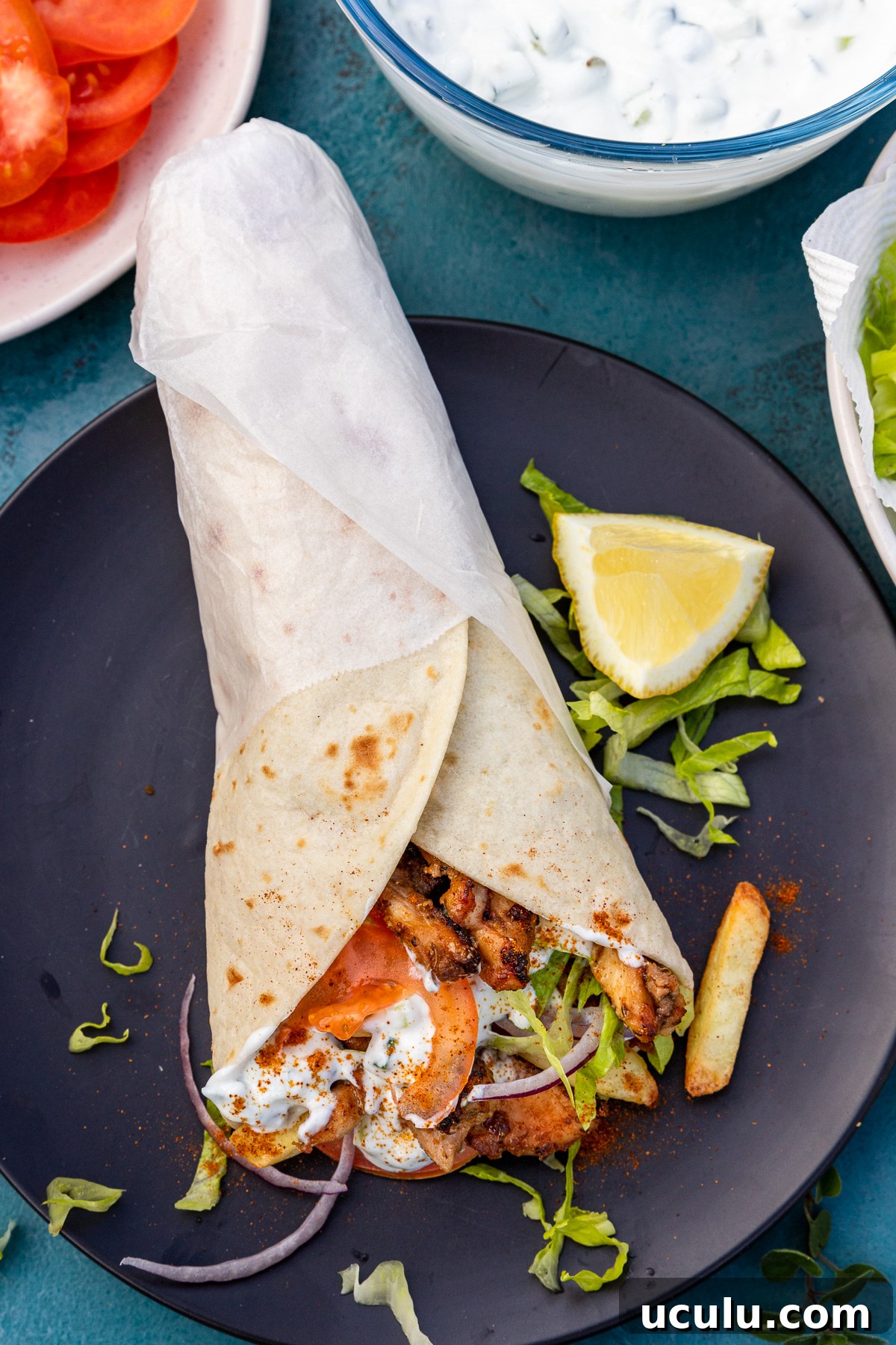 An overhead view of a Greek chicken gryo wrap on a black plate and blue table with tzatziki and other ingredients next to it.