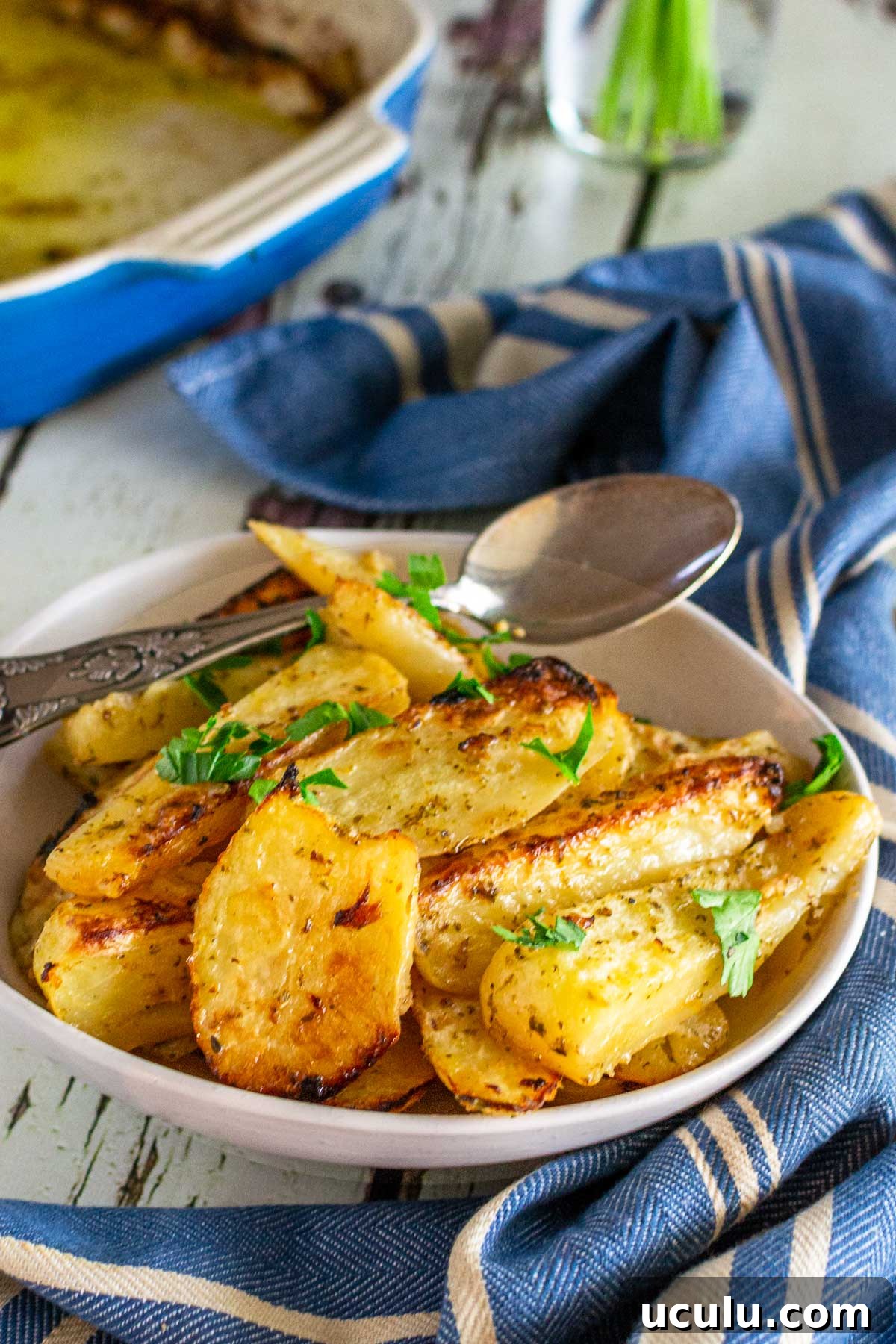 A small dish of Greek potatoes in a beige dish with a spoon in and parsley on top, with a blue and white striped tea towel in front and a blue baking dish at the back, all on a white wooden background.