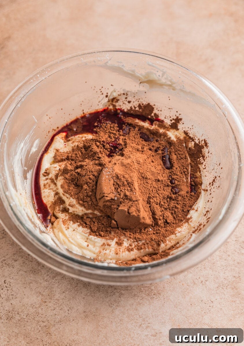 Cocoa powder, vanilla and red food coloring added to creamed cream cheese in glass bowl.