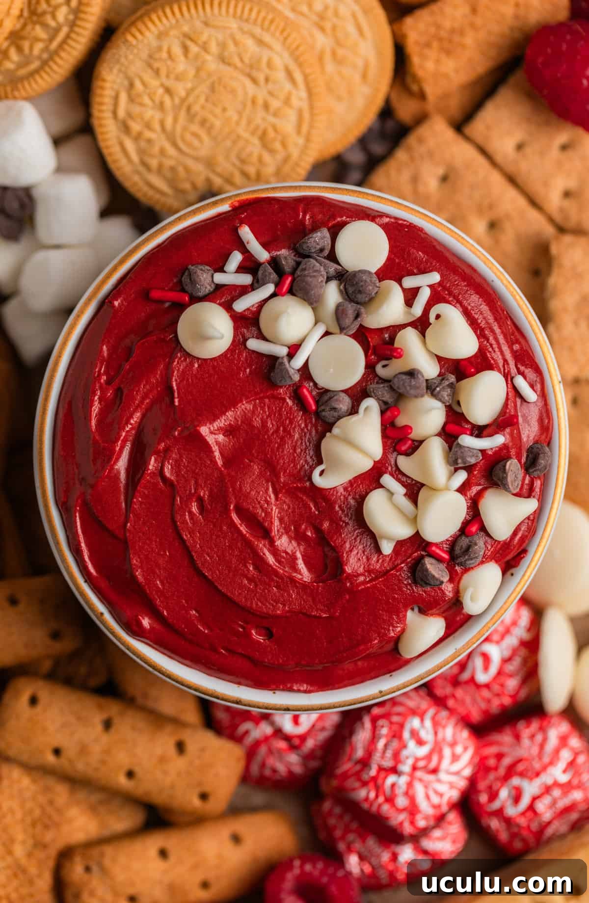 Red velvet cheesecake dip in bowl topped with sprinkles and chocolate chips surrounded by cookies, graham crackers and other dippers.