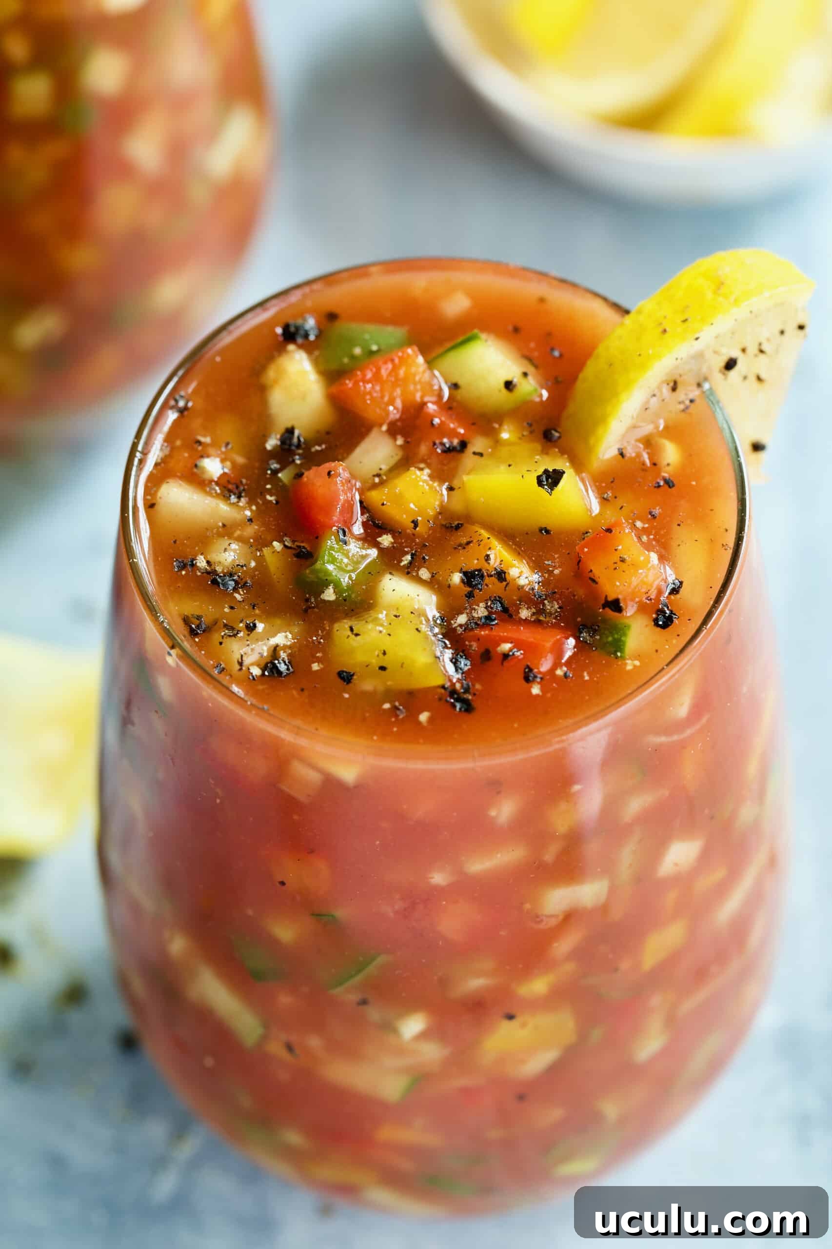 One glass of Best Vegetable Gazpacho on light blue background garnished with lemon wedge.