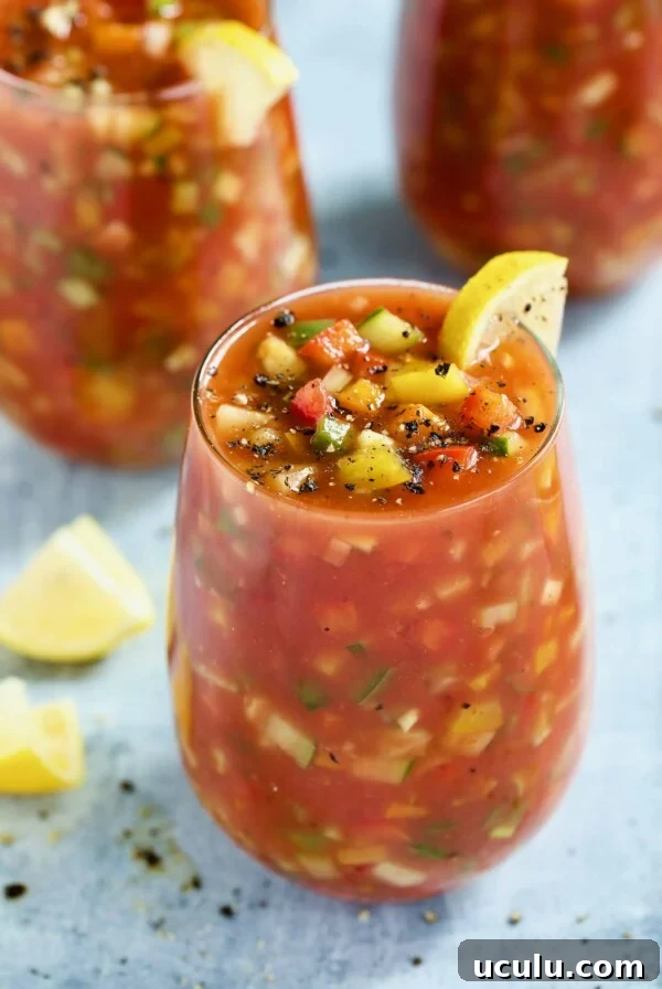 Glass of Very Veggie Gazpacho on light blue background garnished with lemon wedges.