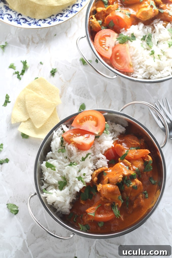 Chicken,coconut & tomato curry served in a metal dish with handles, alongside fluffy white rice and two slices of fresh tomato.