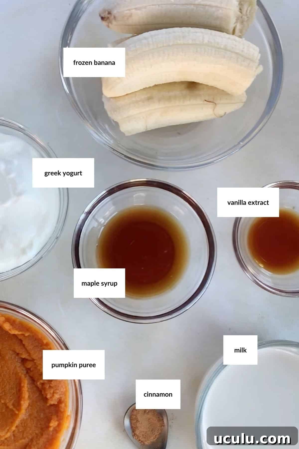 Bowls containing frozen banana, Greek yogurt, maple syrup, vanilla extract, pumpkin puree, cinnamon, and milk are arranged on a white surface, ready to be blended into a delicious pumpkin smoothie.
