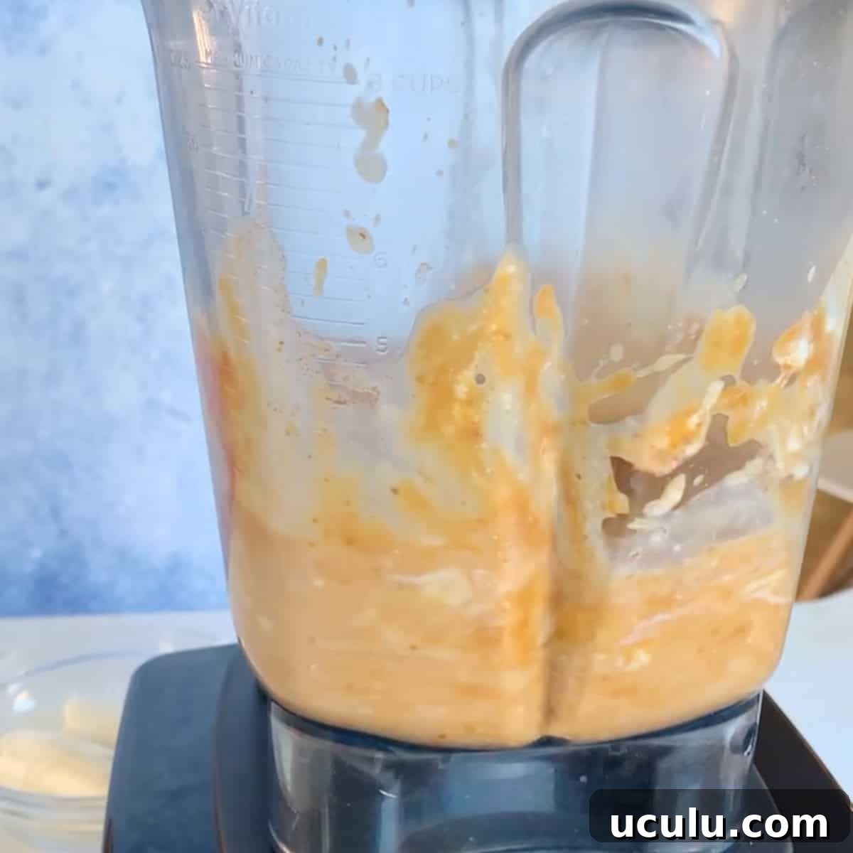 A close-up of a blender mixing an orange and creamy mixture, with some splatters on the sides of the pitcher.