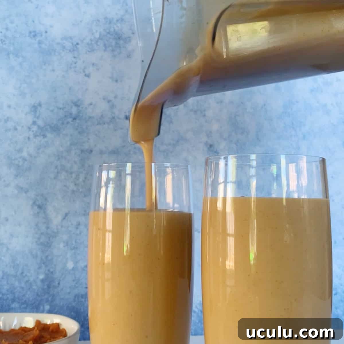 A blender is pouring a thick, light brown smoothie into a glass next to another filled glass, with a small bowl of food in the background.