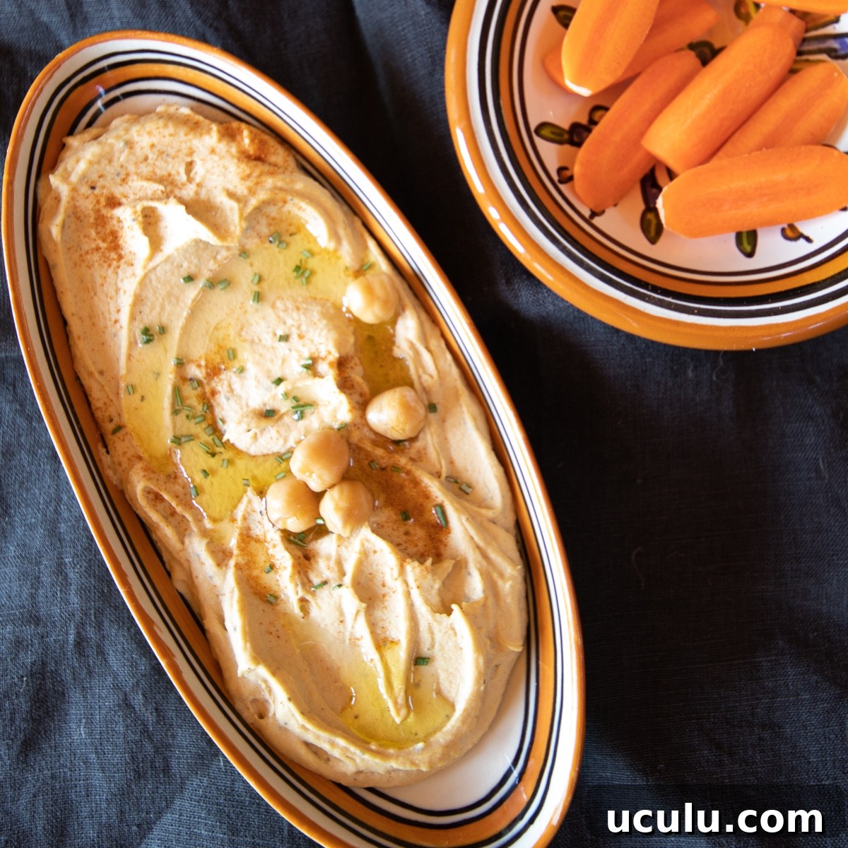 Small dish of creamy smooth hummus with a sprinkling of paprika, and a few chickpeas. Some raw carrots are on the side to dip into the hummus.