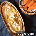 Small dish of creamy smooth hummus with a sprinkling of paprika, and a few chickpeas. Some raw carrots are on the side to dip into the hummus.