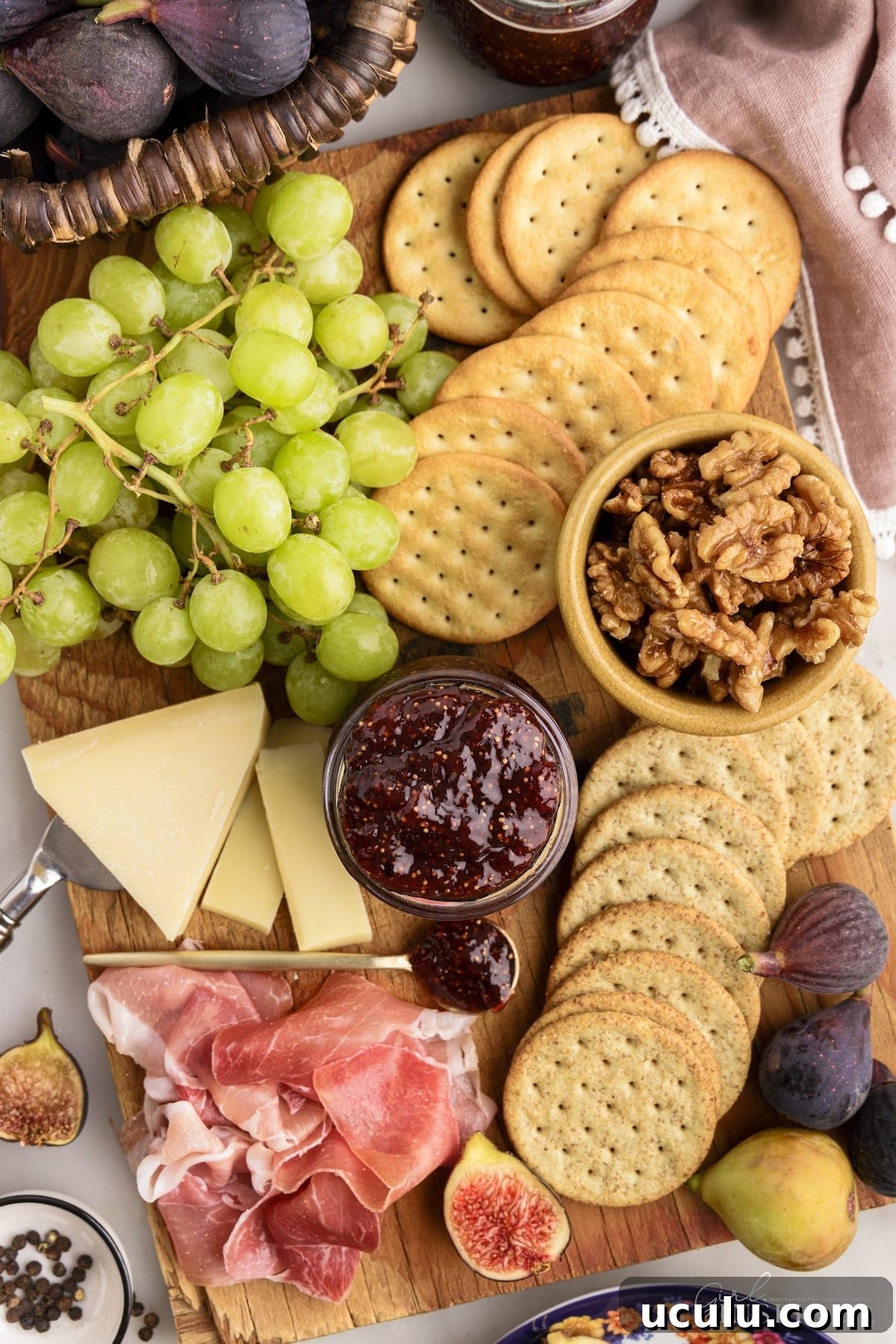 Overhead view of balsamic fig jam in the center of a board, surrounded by crackers, fresh fruit, cheese, and deli meats to enjoy as a snacks platter.