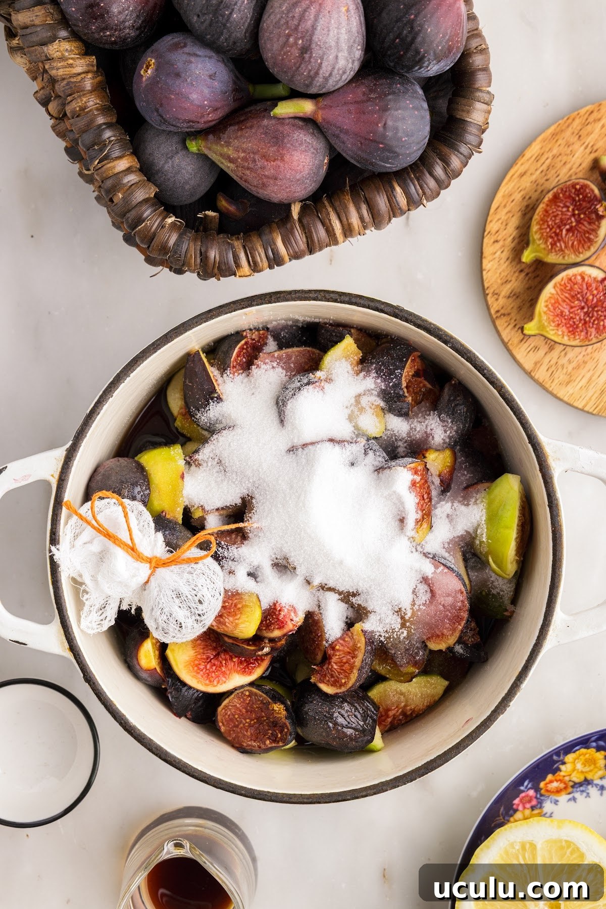 Saucepan with fresh cut figs, sugar, balsamic vinegar, lemon juice, and bundle of peppercorns. Ingredients for this jam recipe in the background.