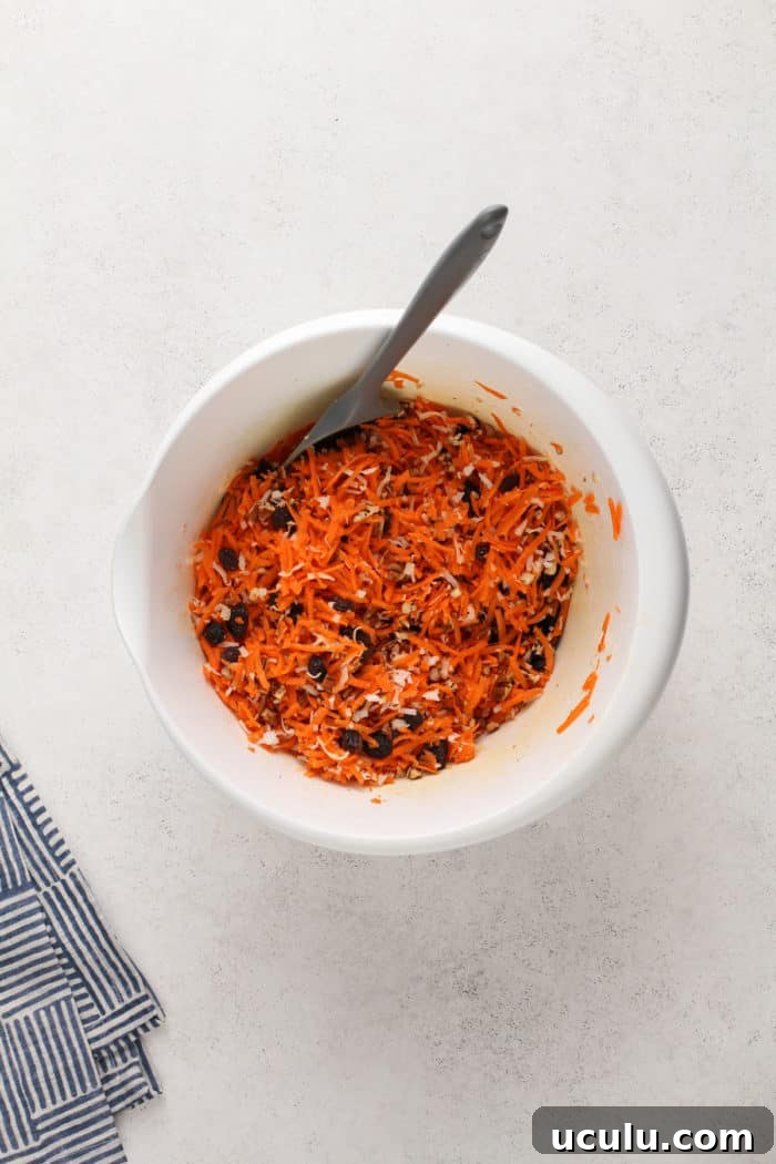 Carrots, nuts, coconut, and raisins mixed in a white mixing bowl.