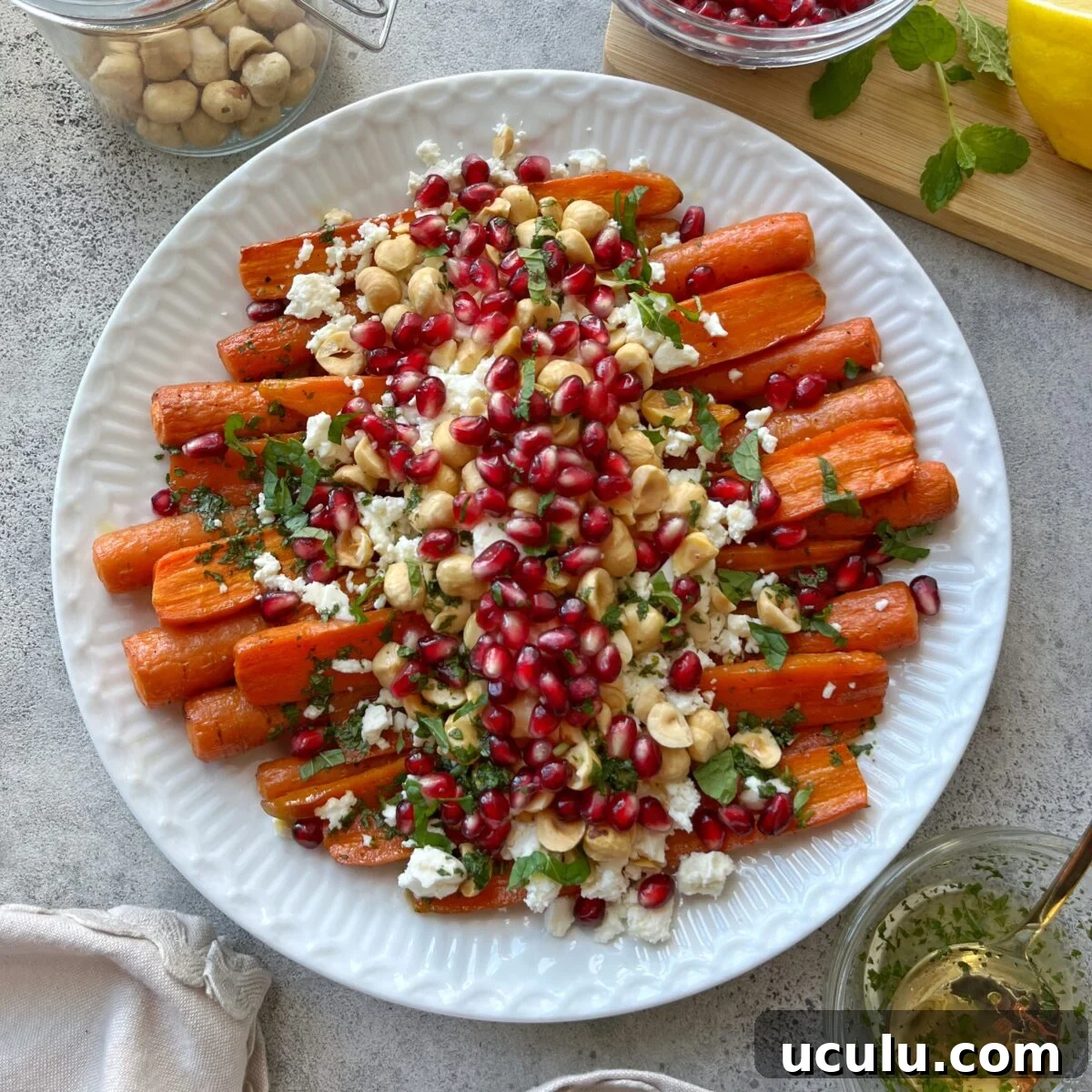A white plate of Roasted Maple Carrots topped with pomegranate seeds, chopped nuts, crumbled cheese, and fresh herbs, with small bowls of ingredients around it.