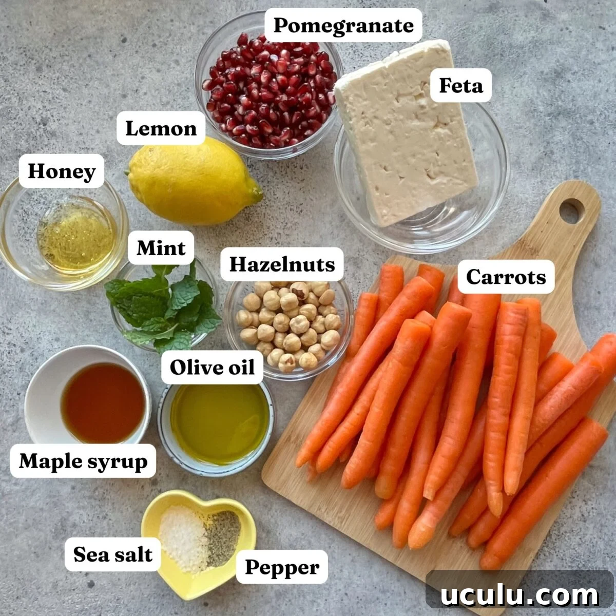 Ingredients for Roasted Maple Carrots are arranged on a surface, including carrots, feta, pomegranate seeds, lemon, honey, mint, hazelnuts, olive oil, maple syrup, sea salt, and pepper.