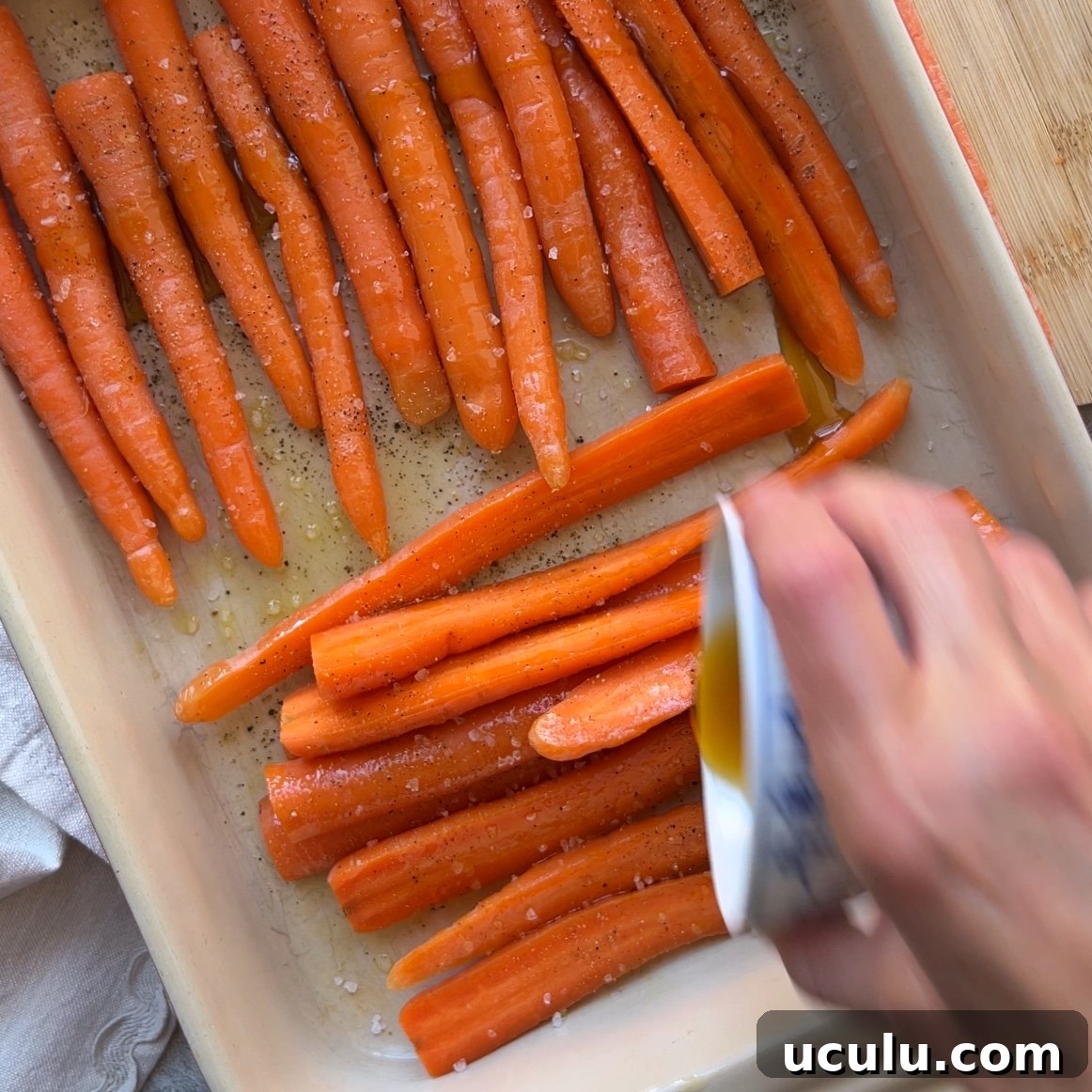 A hand pours maple syrup over raw carrots arranged in a baking dish, sprinkled with salt and pepper.