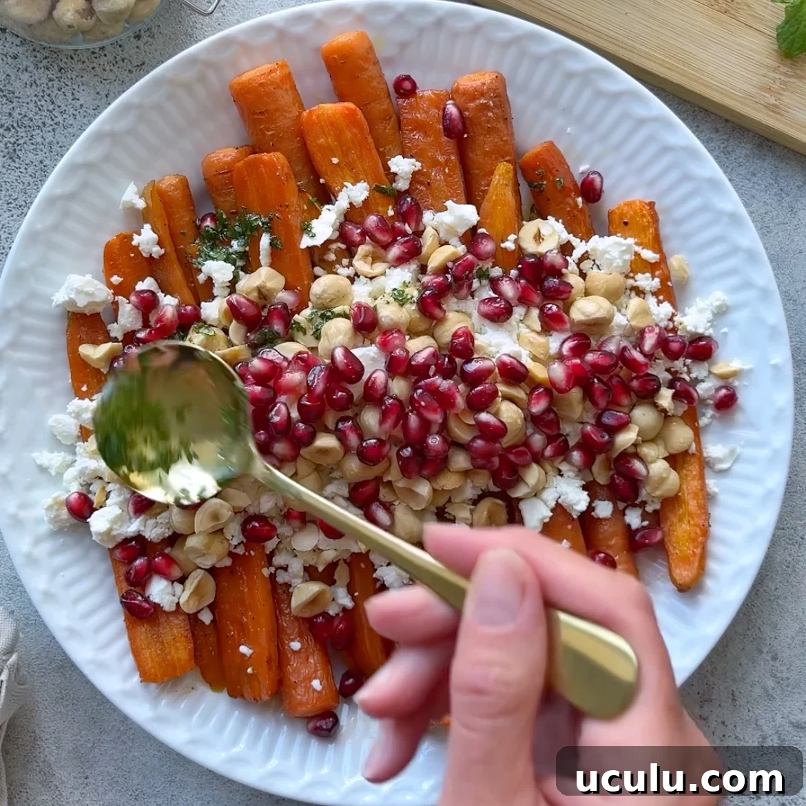 A hand spoons dressing over a plate of Roasted Maple Carrots, topped with feta cheese, hazelnuts, and pomegranate seeds.