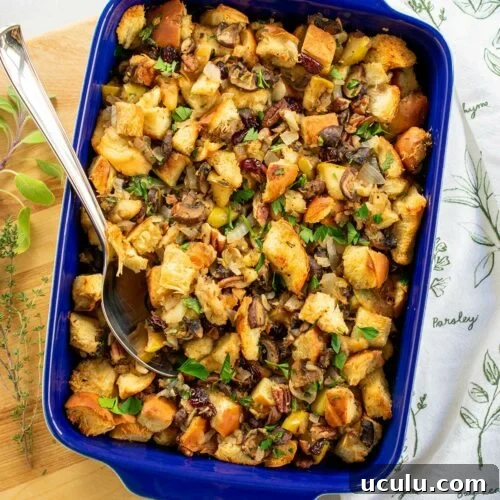 Mushroom challah stuffing in a blue casserole dish on a wooden board with a spoon in the stuffing.