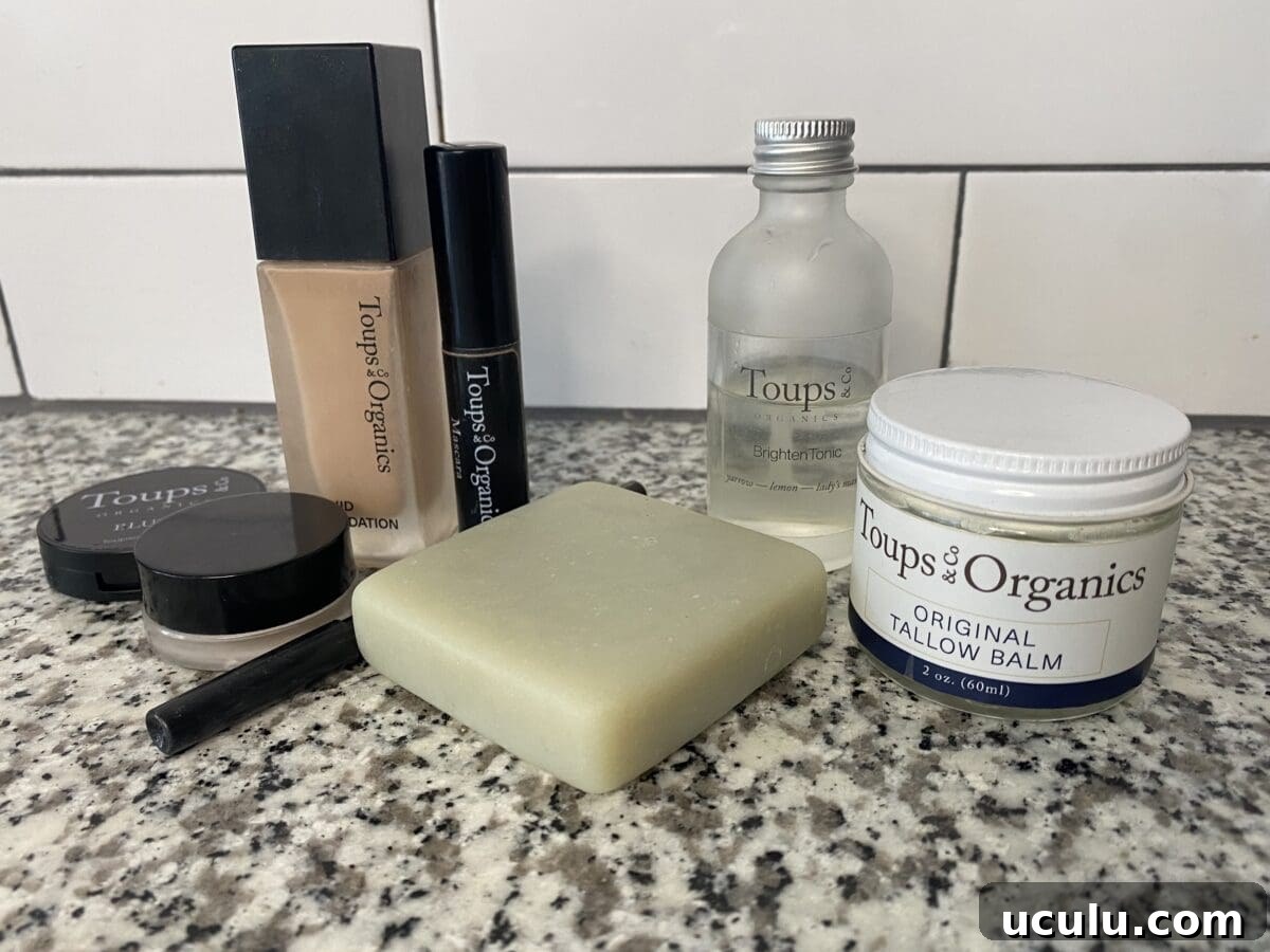 Collection of Toups & Co Organics skincare and makeup products, including liquid foundation, mascara, brightening tonic, tallow balm, soap bar, and small cosmetic containers on a granite counter.