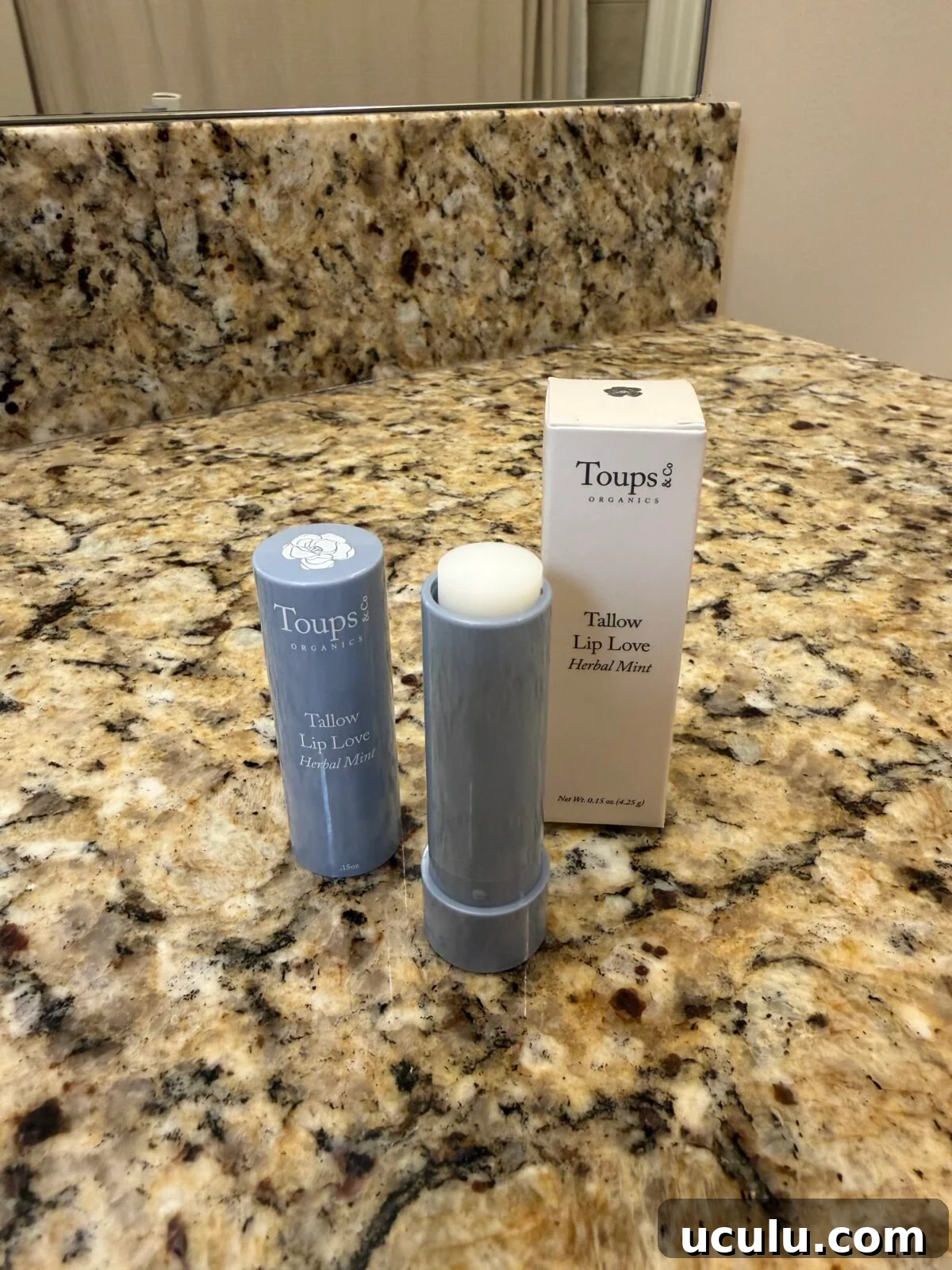 A tube of Toups & Co. Tallow Lip Love Herbal Mint lip balm stands open on a granite bathroom countertop next to its cap and packaging box.
