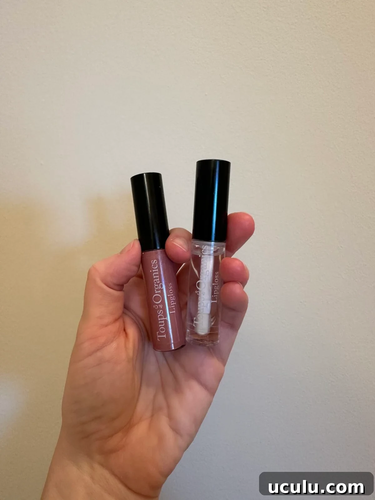 A hand holds two tubes of Tropic Organics lip products, one with a pink shade and the other clear, both with black caps, against a plain beige background.