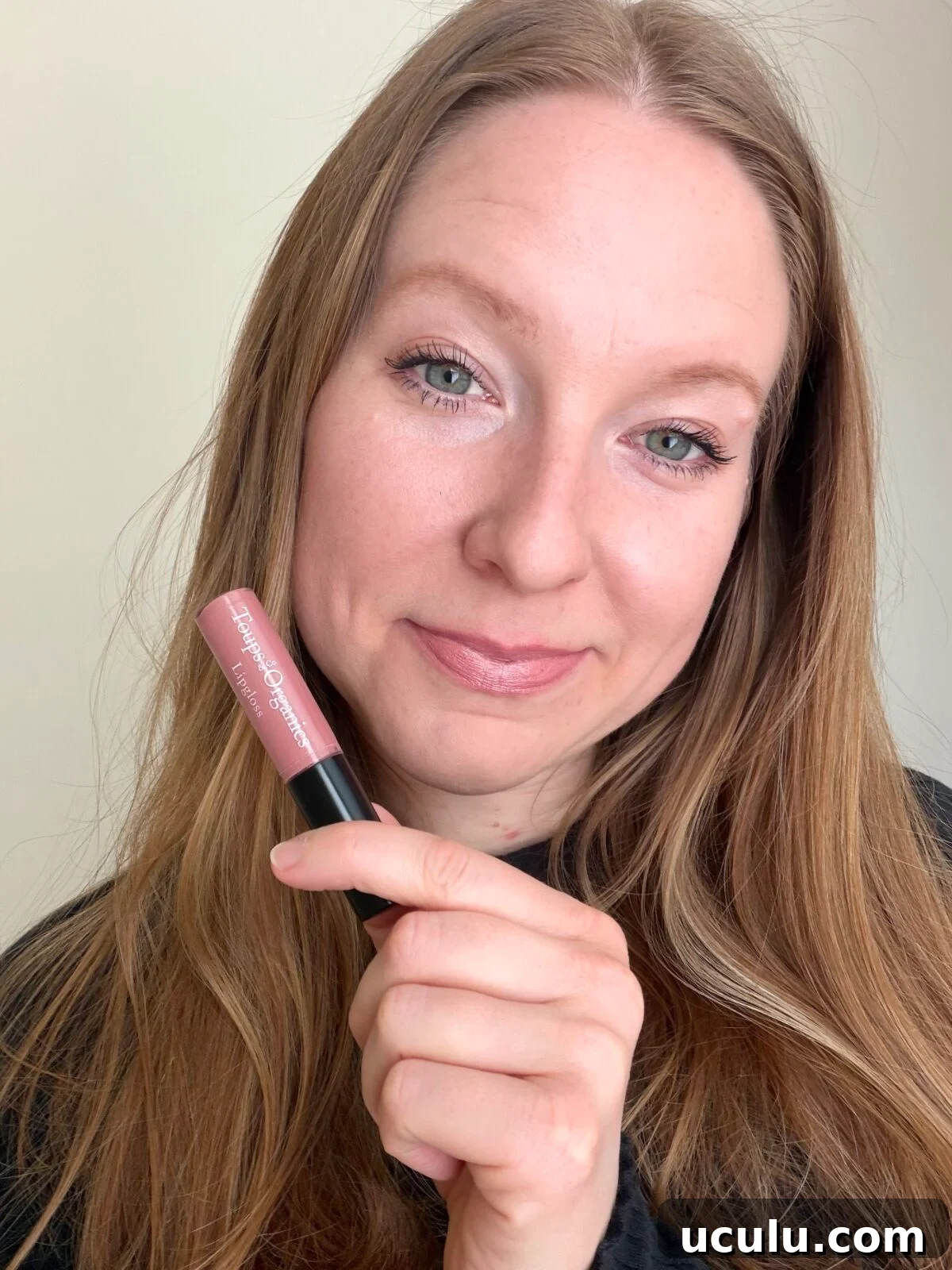 The photo shows a smiling woman with long brown hair holding a tube of pink lip color from the brand Toups & Co Organics in front of her face.