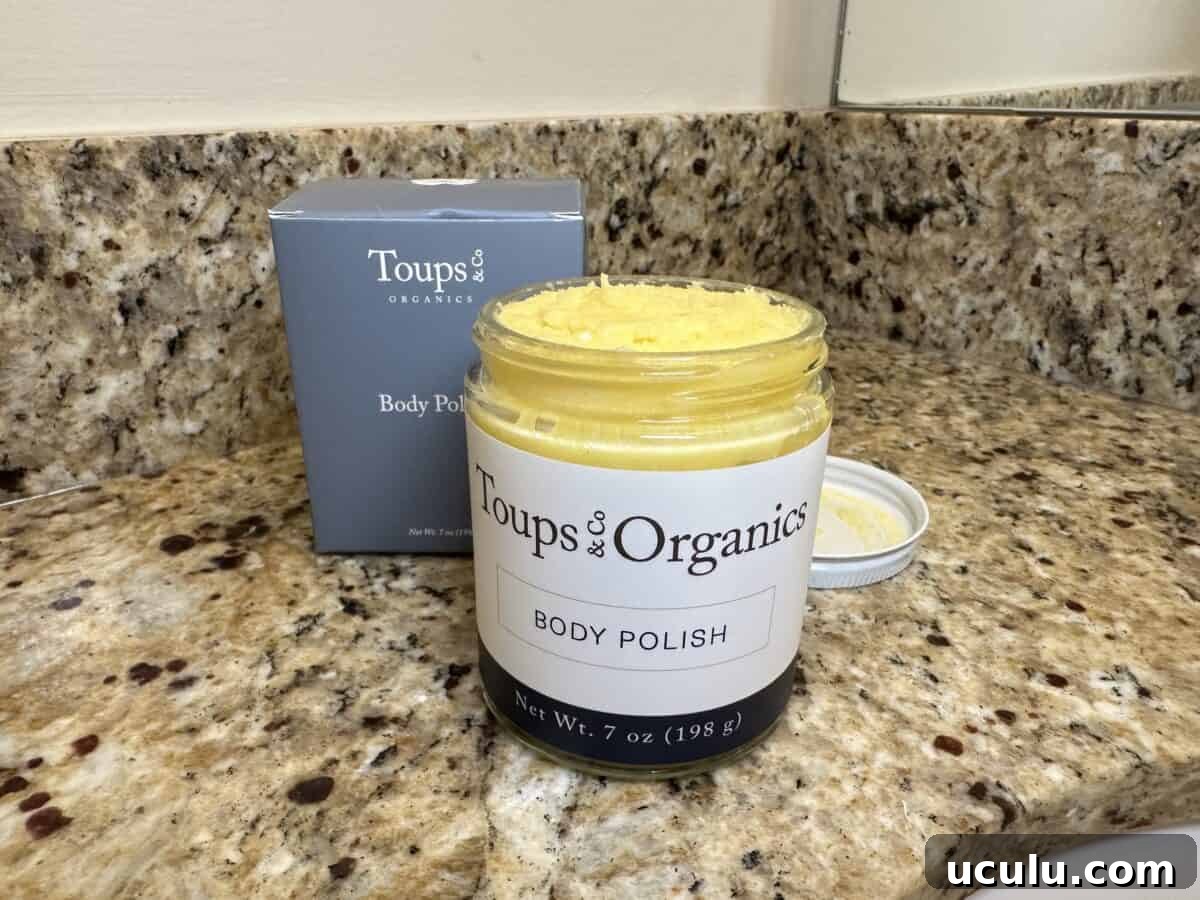 Open jar of Toups & Co Organics Body Polish with yellow scrub inside, sitting on a granite bathroom counter next to its gray product box.