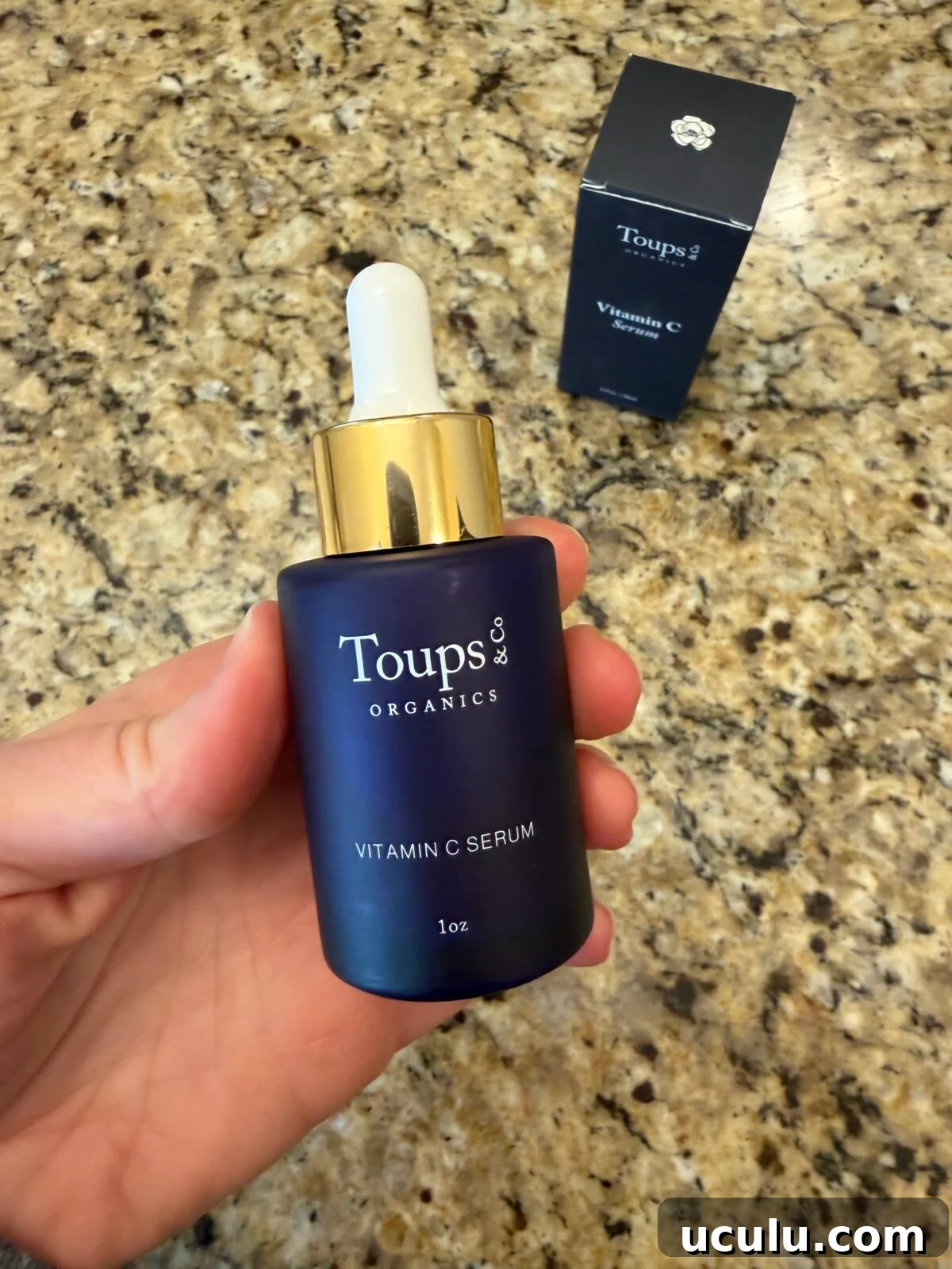 Hand holding Toups & Co Organics Vitamin C Serum bottle with gold dropper cap and white rubber bulb, navy box visible behind