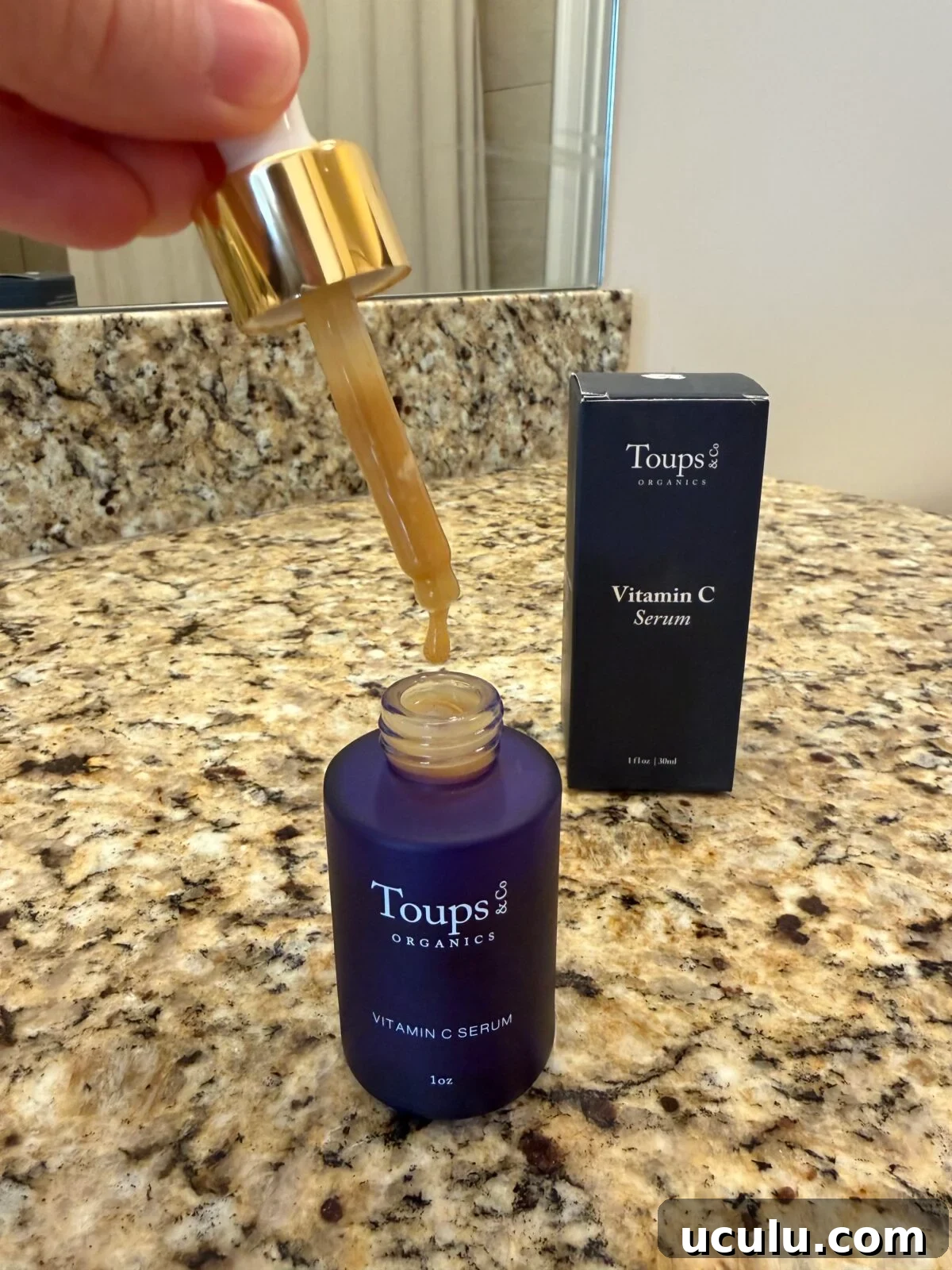 Toups & Co Organics Vitamin C Serum bottle with dropper dispensing golden serum, navy box in background on granite counter