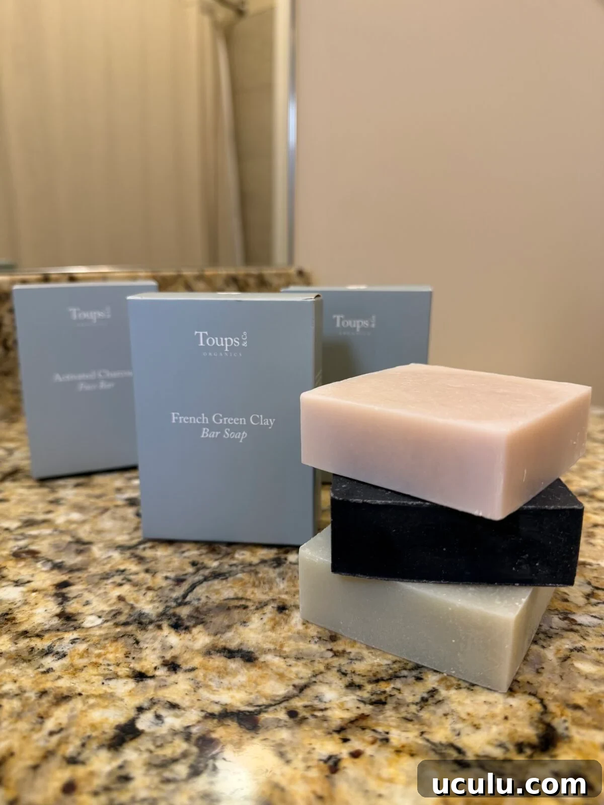 Three bar soaps—one light pink, one black, and one light green—are stacked on a granite countertop.