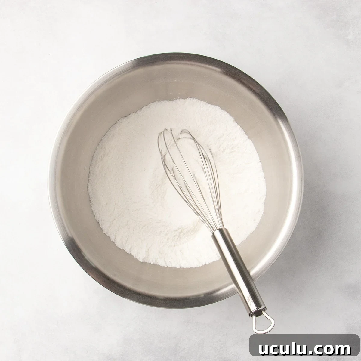 Metal mixing bowl with white dry ingredients and a whisk resting inside.